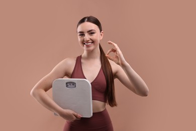 Happy woman with floor scale showing ok gesture on beige background