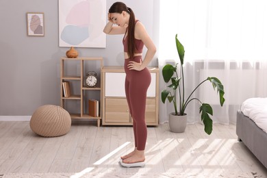 Photo of Worried woman standing on floor scale at home