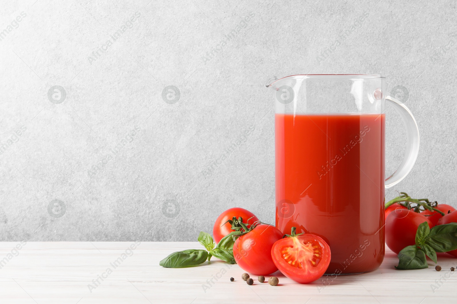 Photo of Tasty tomato juice in jug, basil, peppercorns and vegetables on white wooden table indoors. Space for text