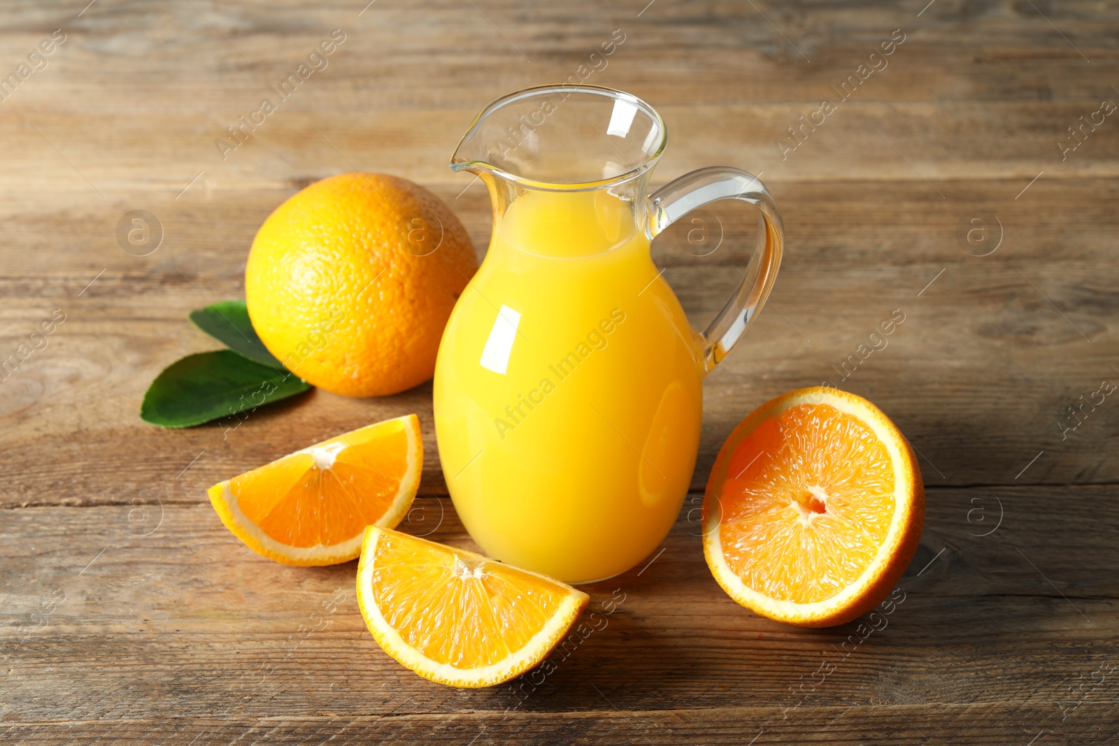 Photo of Tasty orange juice in glass jug, green leaves and fresh fruits on wooden table