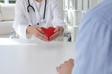 Doctor showing red heart to patient at white table in clinic, closeup