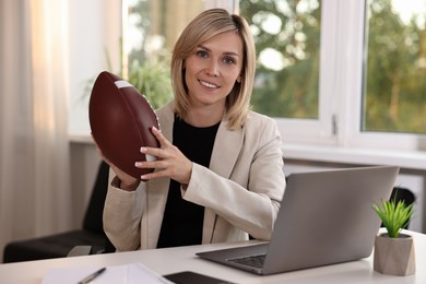 Photo of Smiling woman with american football ball at table in office