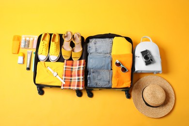 Open suitcase with traveler's belongings on yellow background, flat lay