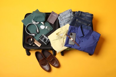 Open suitcase with traveler's belongings on yellow background, flat lay