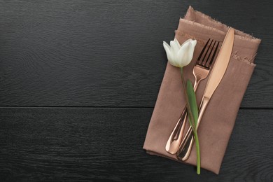 Stylish setting with cutlery, flower and napkin on black wooden table, top view. Space for text