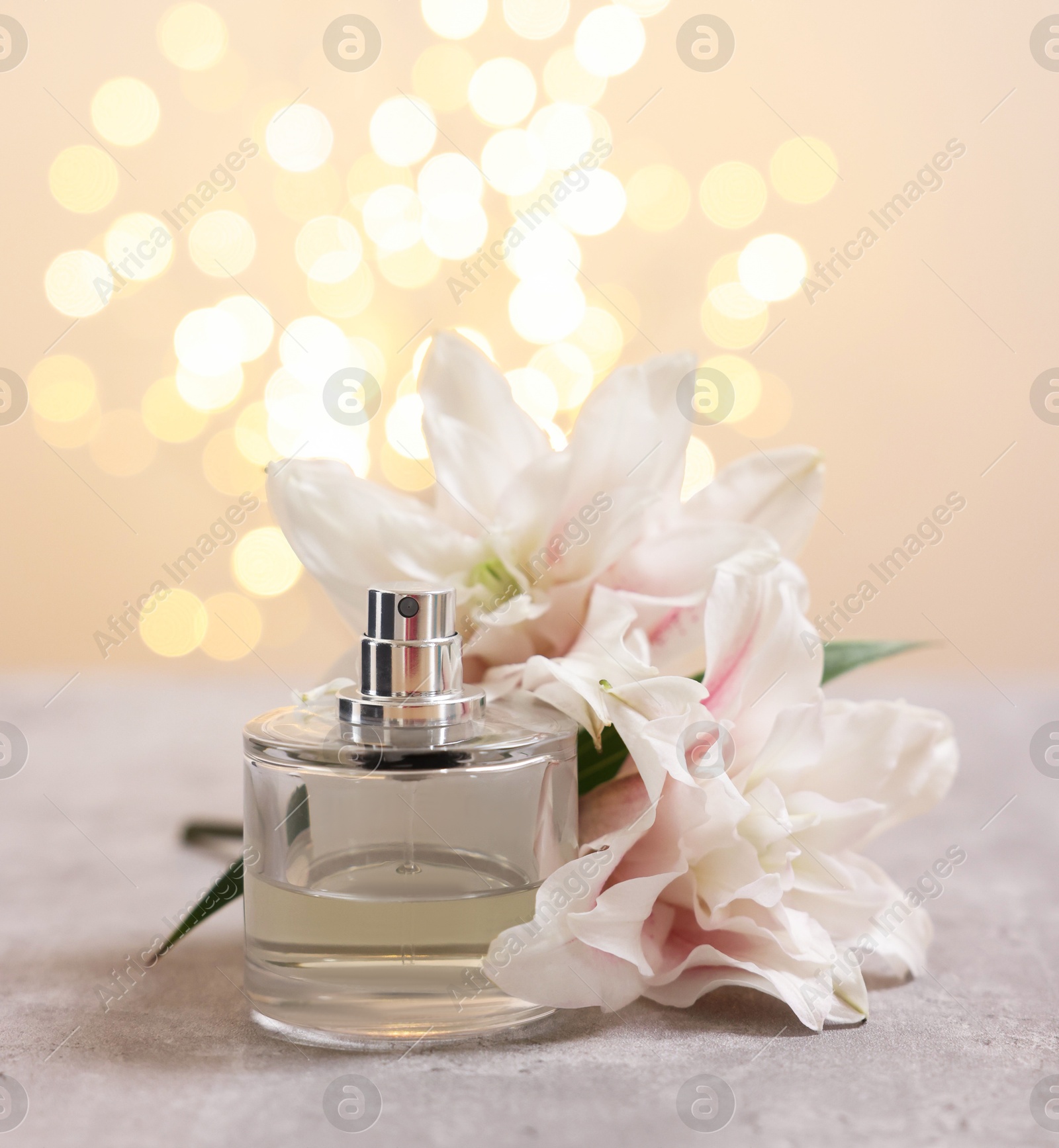 Photo of Bottle of perfume and beautiful lily flowers on table against beige background with blurred lights, space for text