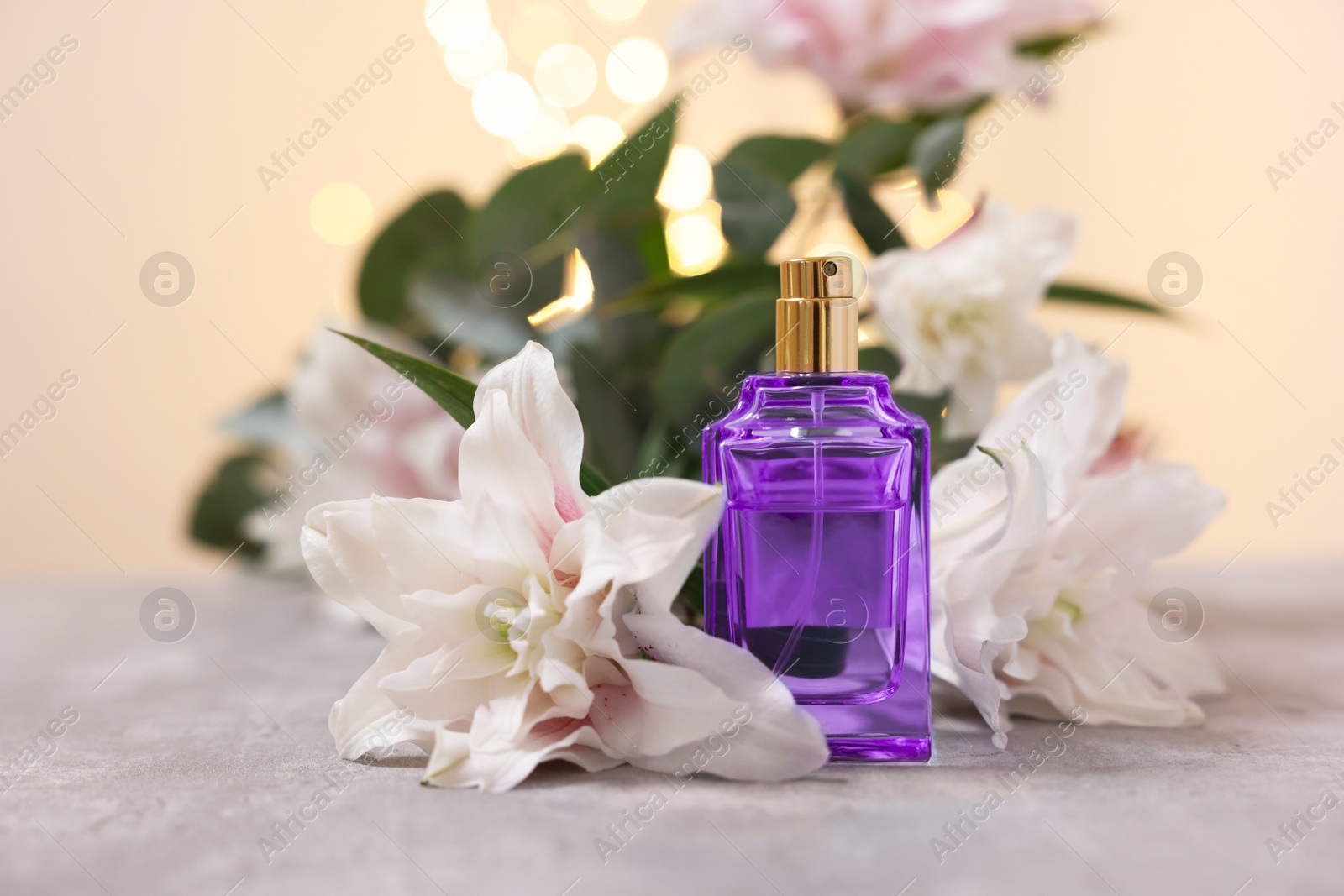 Photo of Bottle of perfume and beautiful lily flowers on table against beige background with blurred lights, closeup