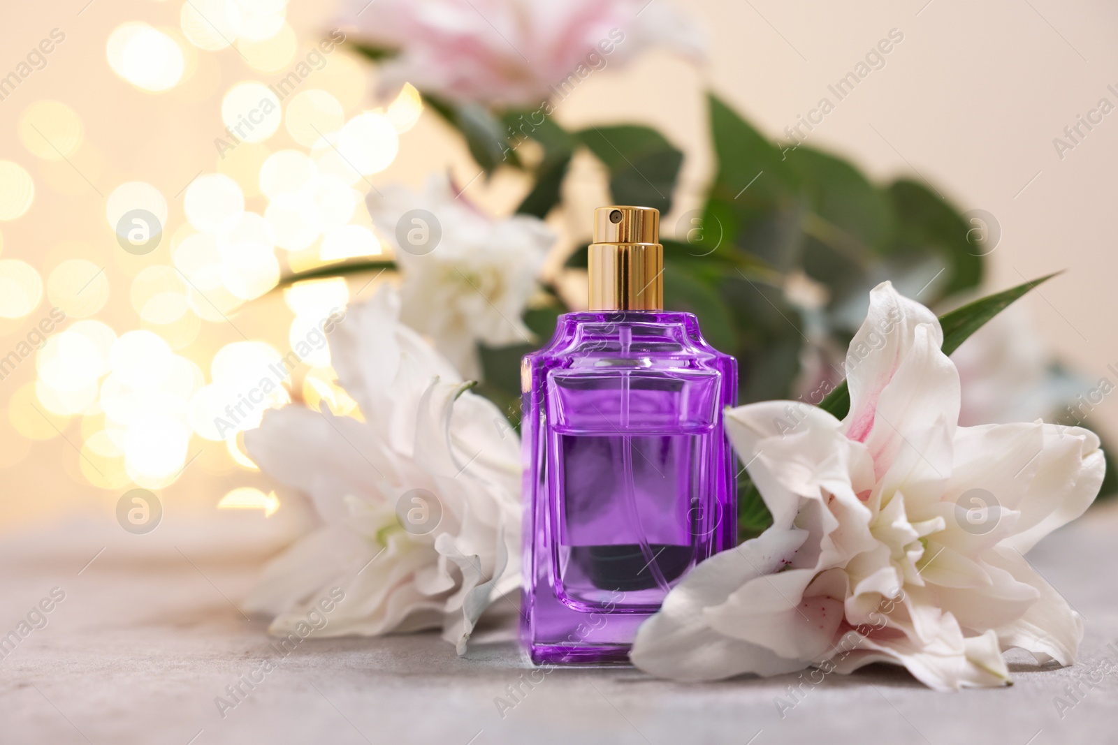 Photo of Bottle of perfume and beautiful lily flowers on table against beige background with blurred lights, closeup