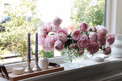 Beautiful pink peonies in vase and tray with candles on window sill. Interior design