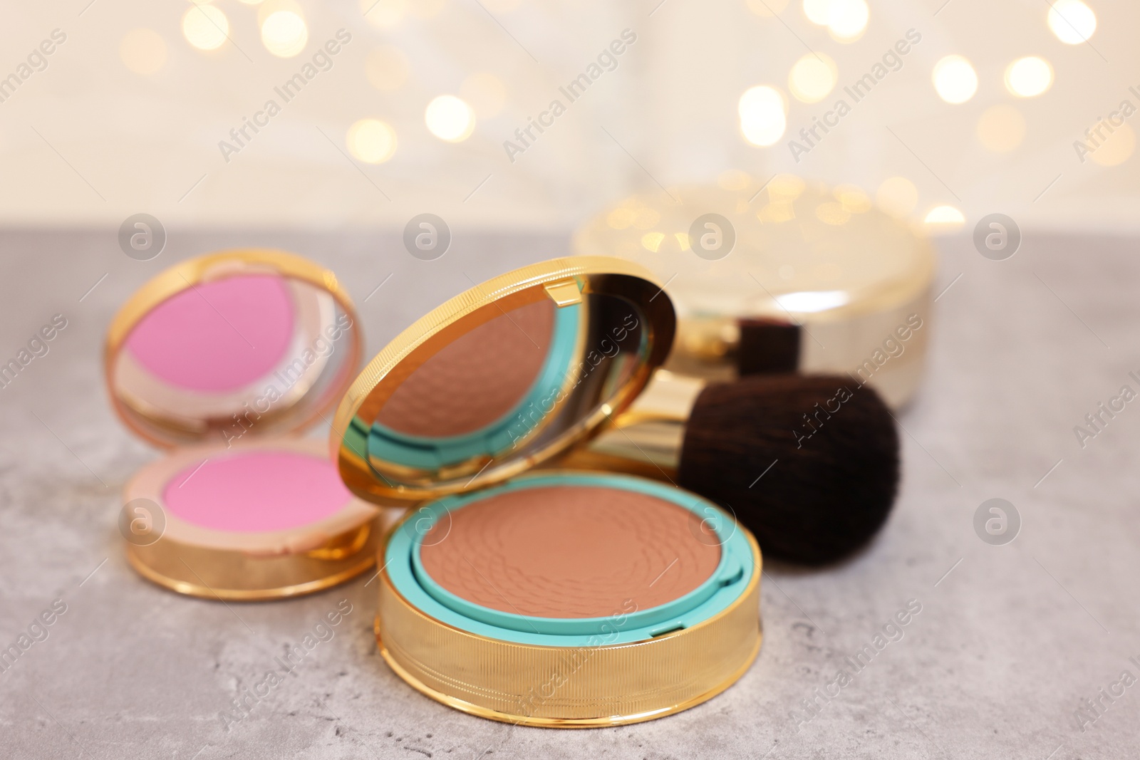 Photo of Face bronzer, blusher and makeup brush on grey textured table against blurred lights, closeup