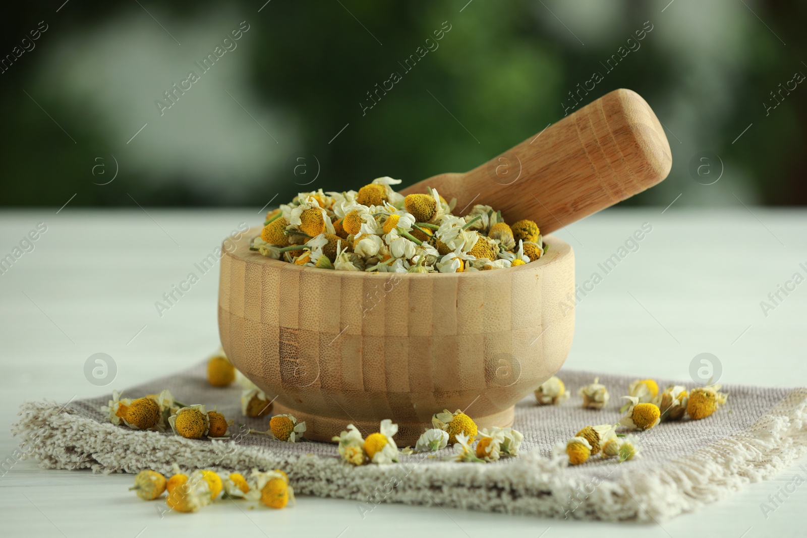 Photo of Mortar and pestle with chamomile flowers on light table against blurred green background