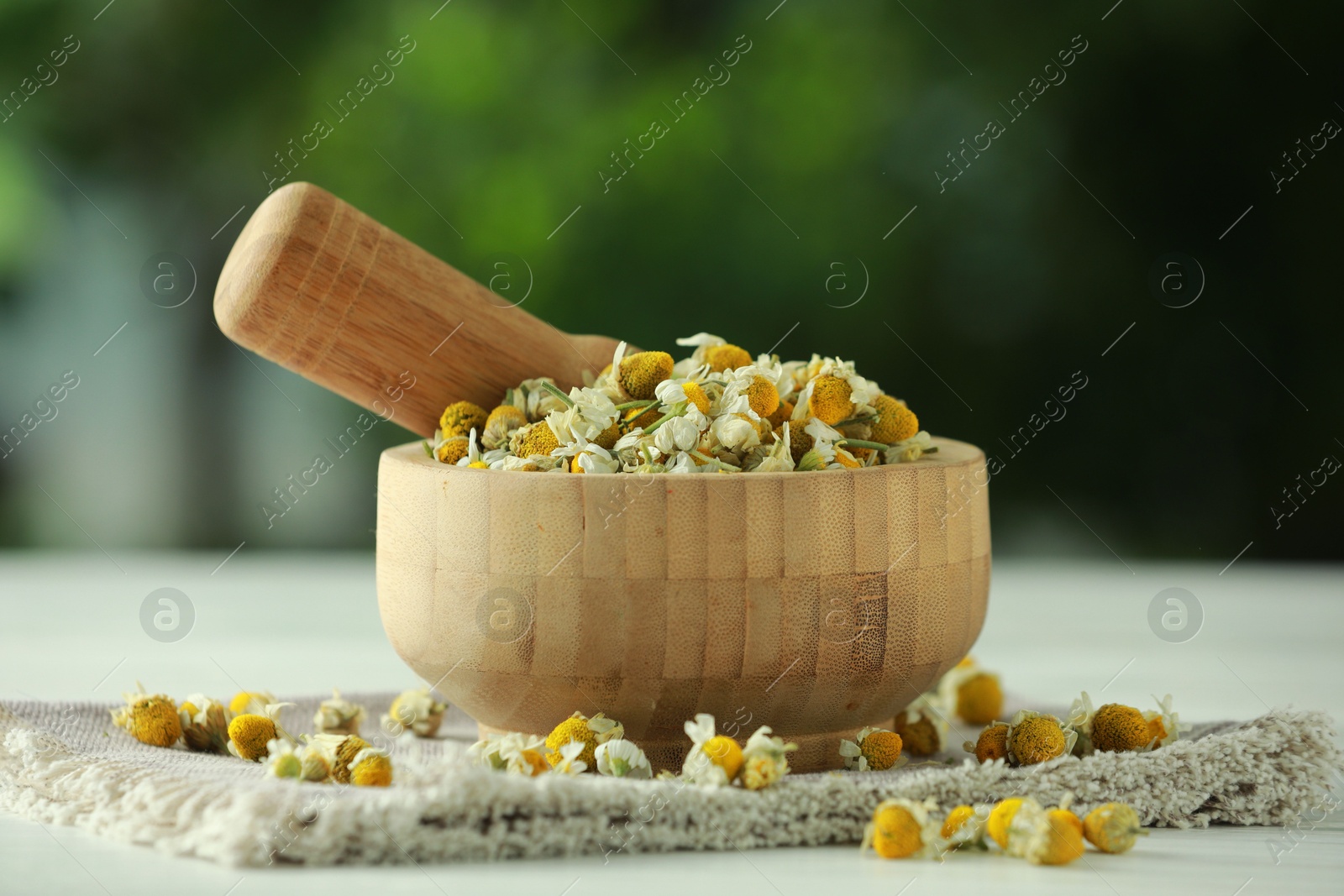 Photo of Mortar and pestle with chamomile flowers on light table against blurred green background