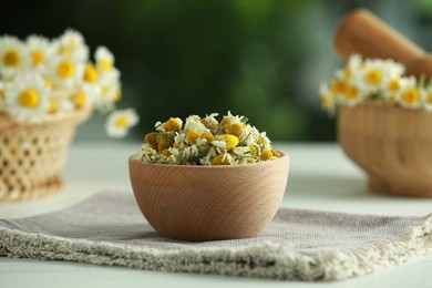 Photo of Chamomile flowers in bowl on light table against blurred green background