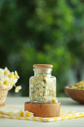 Photo of Dry and fresh chamomile flowers in glass jar on light table against blurred green background, space for text