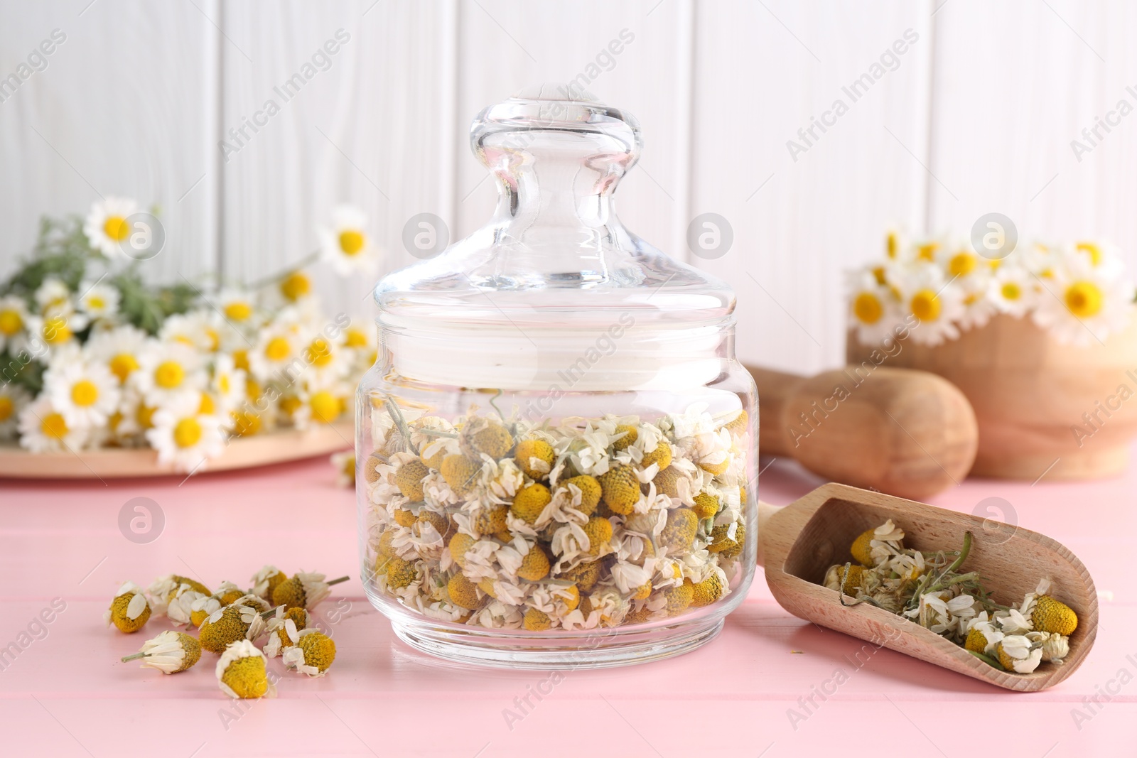 Photo of Chamomile flowers in glass jar and scoop on pink wooden table