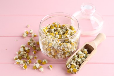 Photo of Chamomile flowers in glass jar and scoop on pink wooden table, closeup