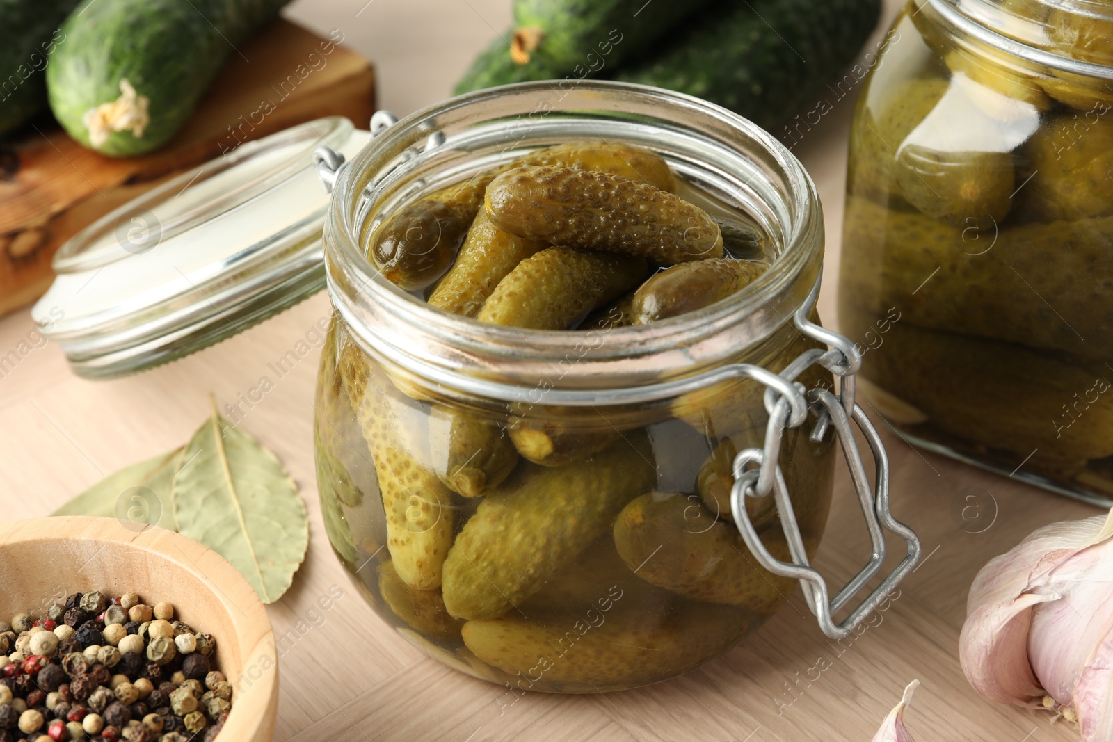 Photo of Pickled cucumbers in open jar, peppercorns and garlic on wooden table, closeup