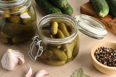 Photo of Pickled cucumbers in jars, peppercorns and garlic on wooden table, closeup