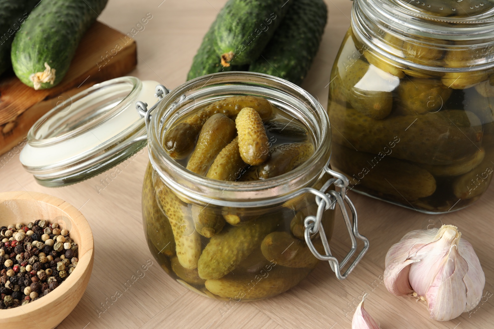 Photo of Pickled cucumbers in jars, peppercorns and garlic on wooden table, closeup