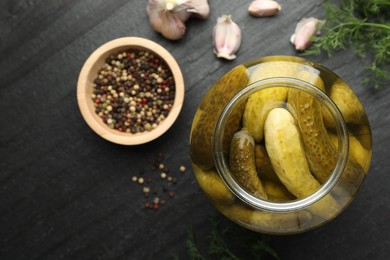 Pickled cucumbers in open jar and spices on dark textured table, flat lay