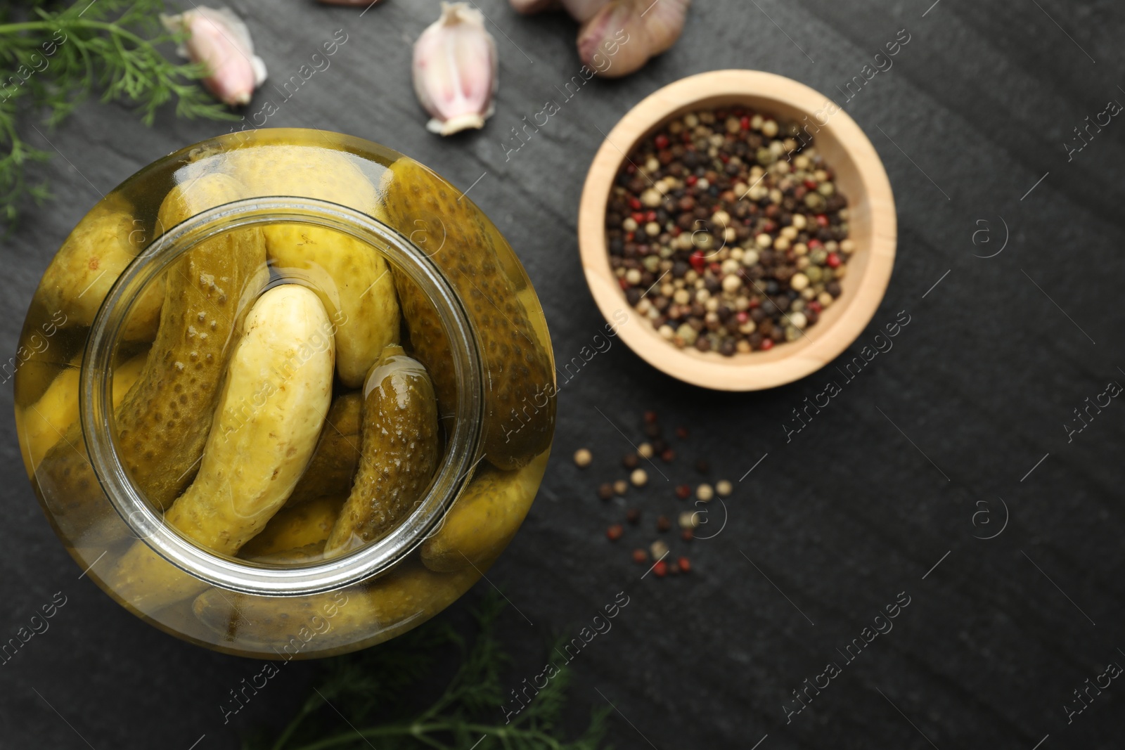 Photo of Pickled cucumbers in open jar and spices on dark textured table, flat lay