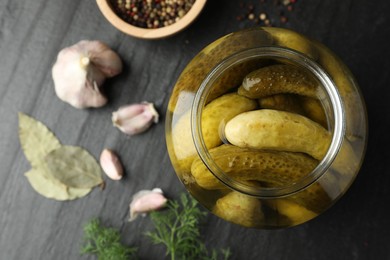 Pickled cucumbers in open jar and spices on dark textured table, flat lay