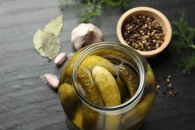 Photo of Pickled cucumbers in open jar and spices on dark textured table, closeup