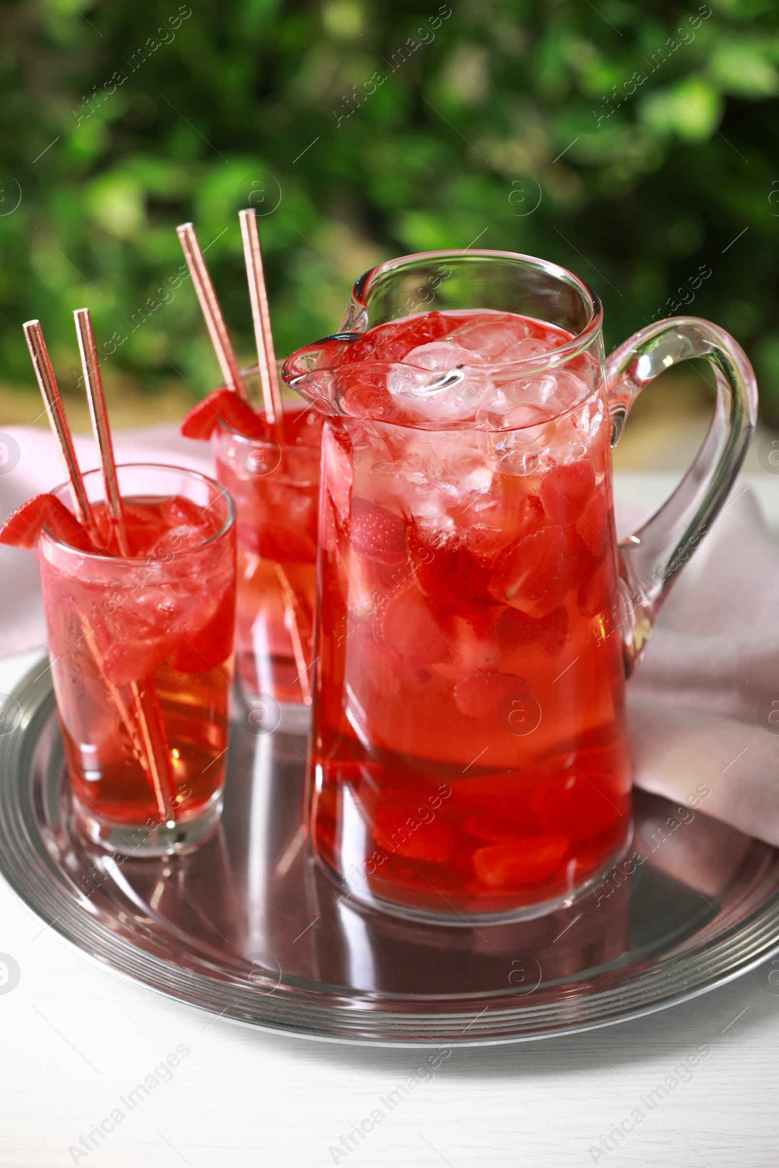 Photo of Tasty strawberry lemonade in jug and glasses on white table against blurred green background