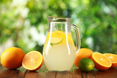 Photo of Refreshing lemonade with citruses in jug among fruits on wooden table against blurred green background