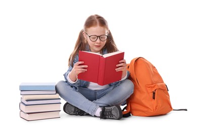 Cute little girl reading book near backpack on white background