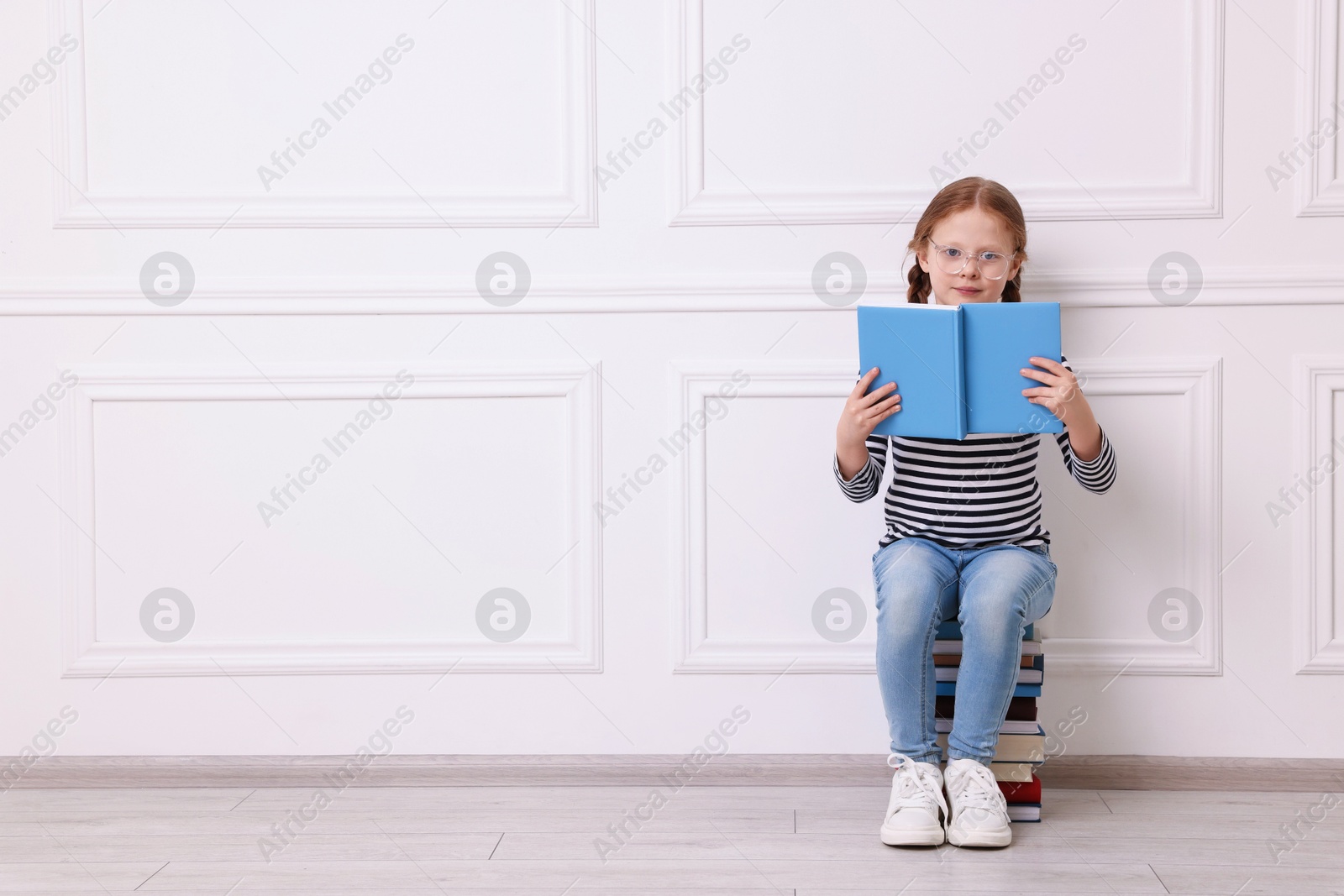 Photo of Cute girl reading while sitting on stack of books indoors. Space for text