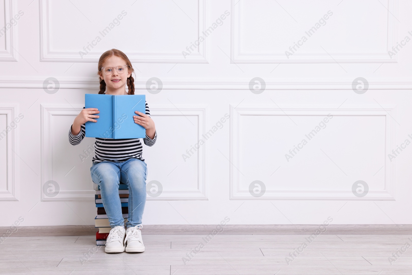 Photo of Smiling girl sitting on stack of books indoors. Space for text