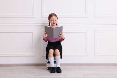 Cute girl reading while sitting on stack of books indoors