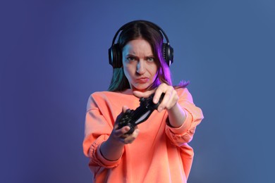 Emotional woman in headphones playing video game with controller on dark blue background