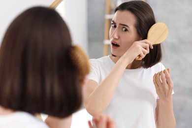 Photo of Emotional woman brushing her hair near mirror indoors. Alopecia problem