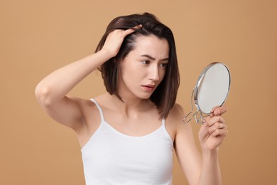 Sad woman with hair loss problem looking at mirror on light brown background