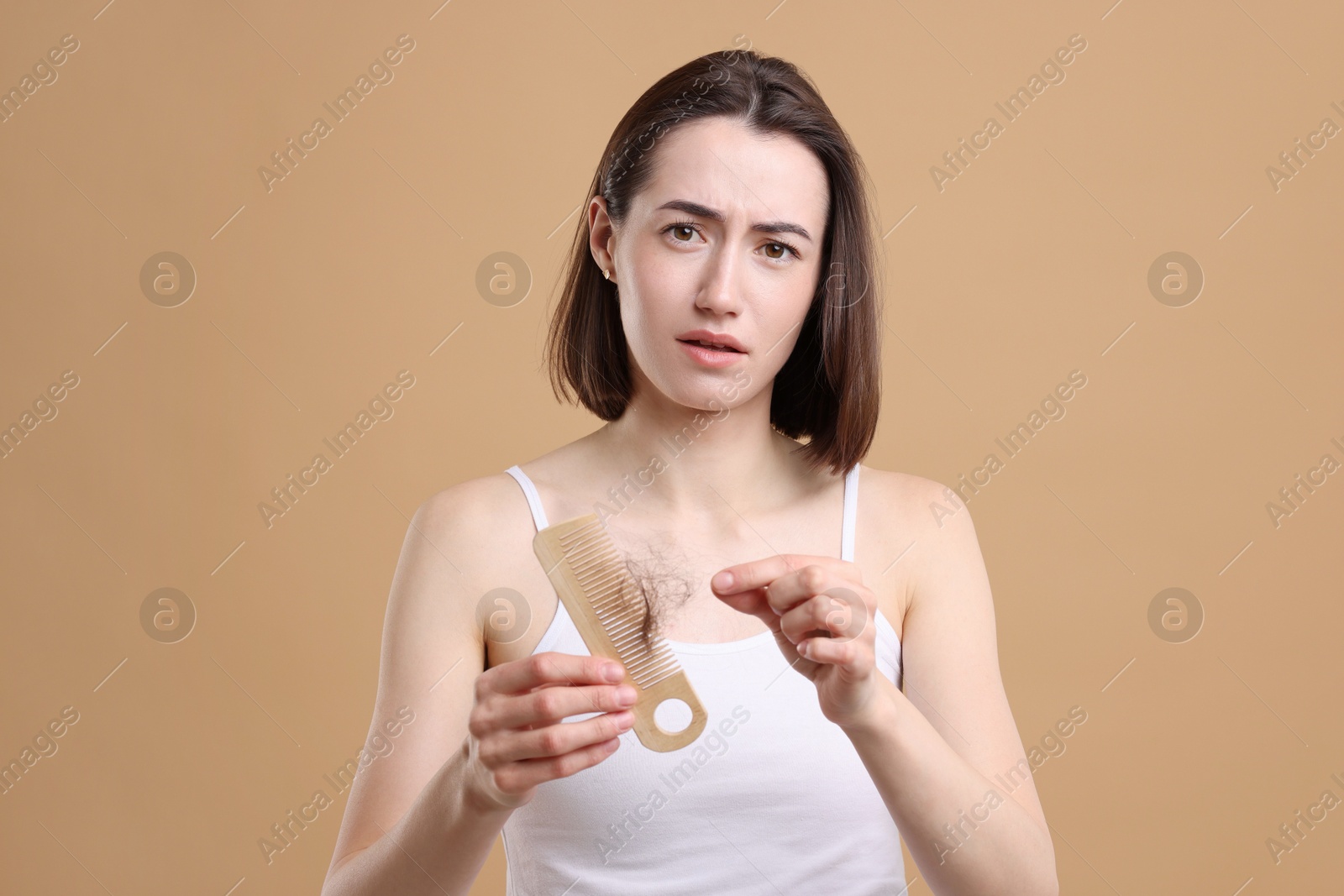 Photo of Sad woman taking her lost hair from comb on light brown background. Alopecia problem
