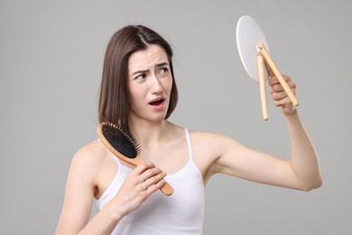 Emotional woman with hair loss problem looking at mirror on grey background