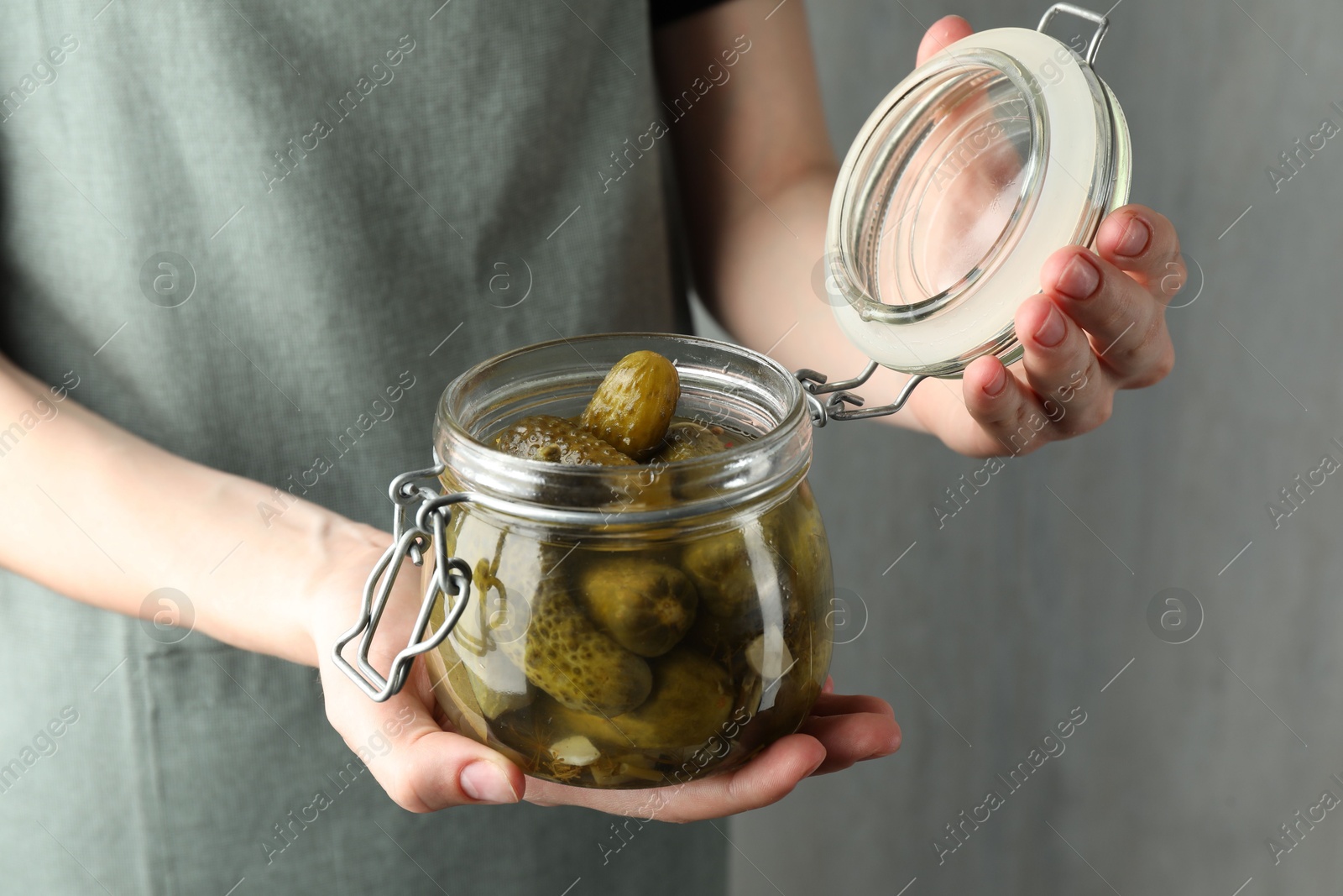 Photo of Woman opening jar with pickled cucumbers on grey background, closeup