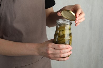 Woman opening jar with pickled cucumbers on grey background, closeup