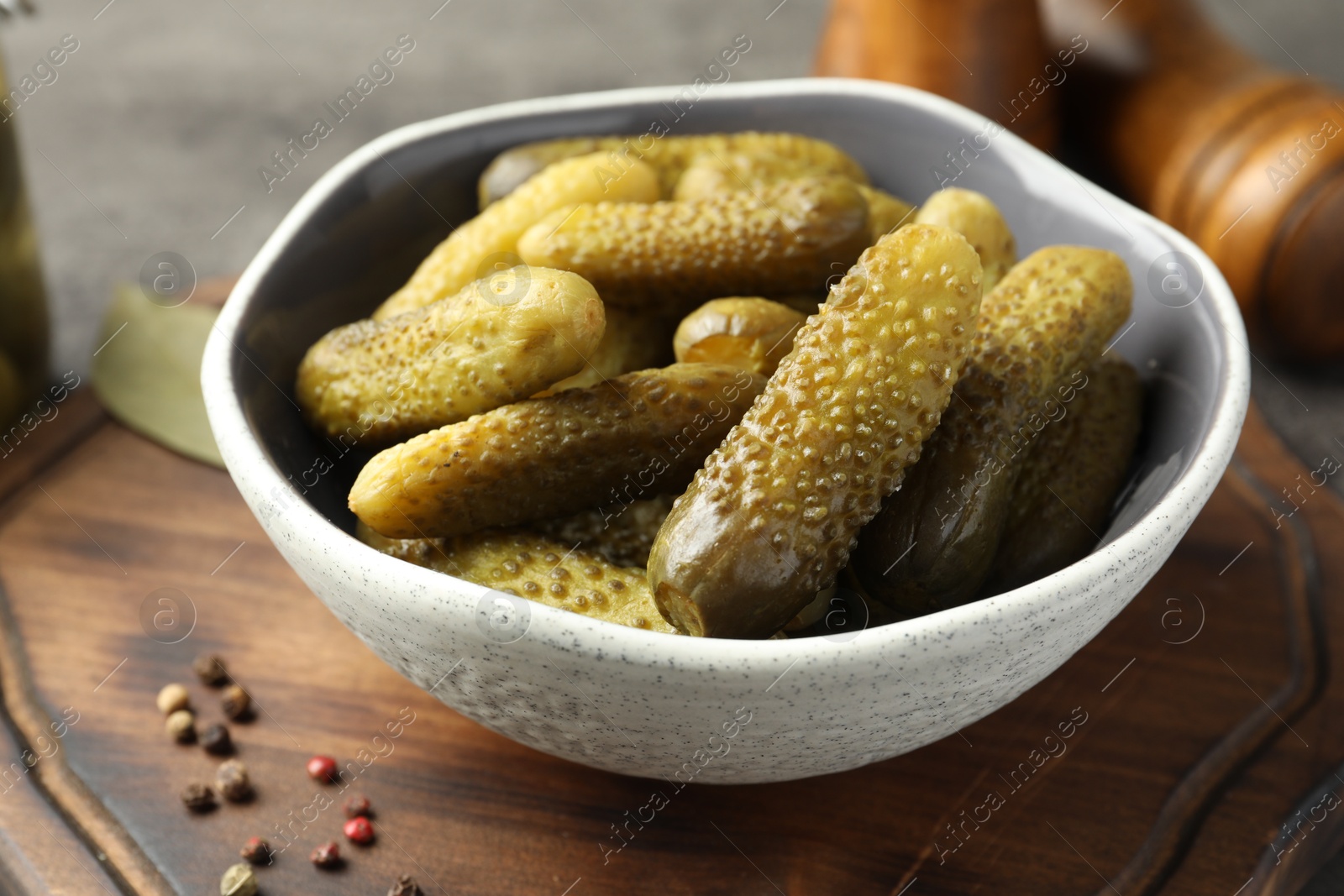 Photo of Pickled cucumbers in bowl and peppercorns on wooden board, closeup