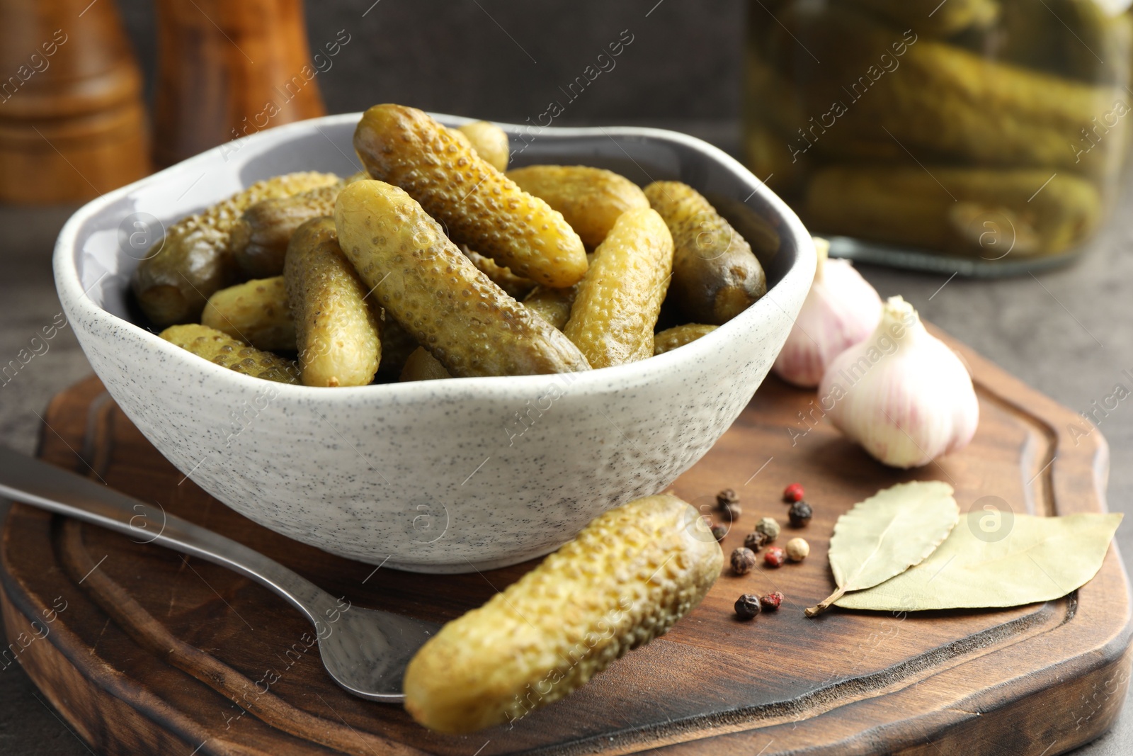 Photo of Pickled cucumbers in bowl, fork, peppercorns, bay leaves and garlic on table, closeup