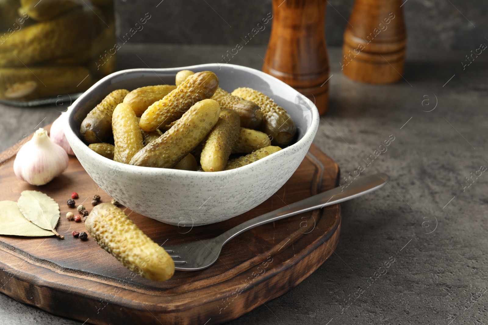 Photo of Pickled cucumbers in bowl, fork, peppercorns, bay leaves and garlic on grey textured table