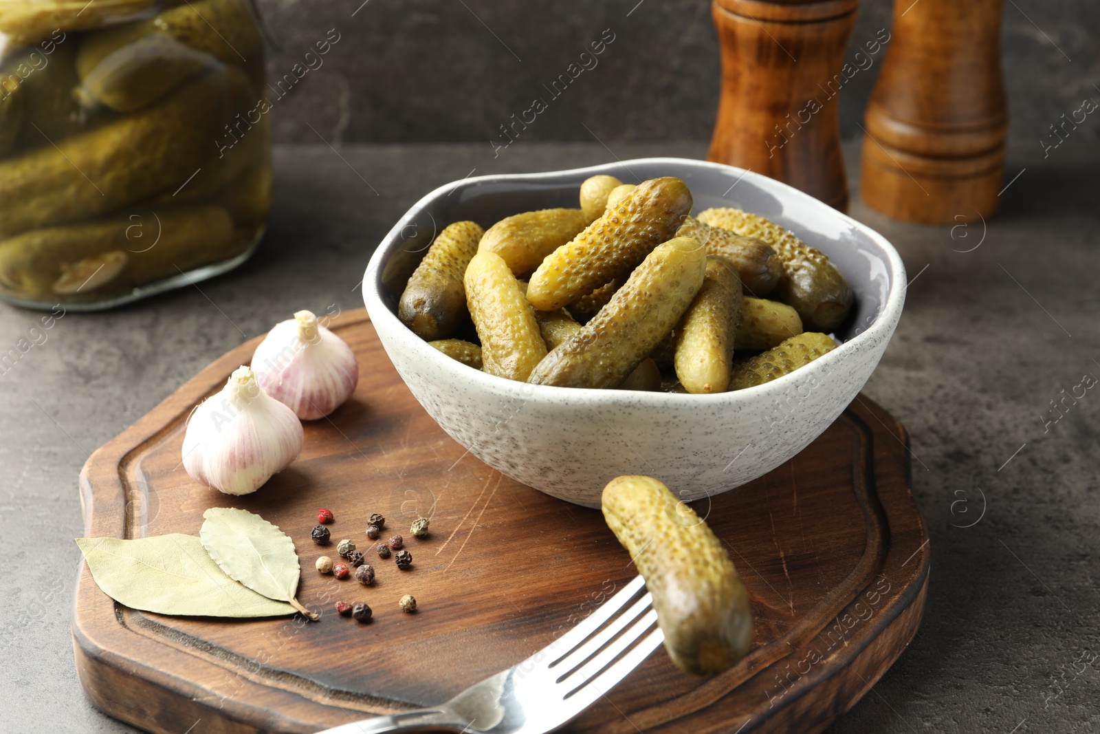 Photo of Pickled cucumbers in bowl, fork, peppercorns, bay leaves and garlic on grey textured table, closeup