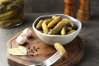 Photo of Pickled cucumbers in bowl, fork, peppercorns, bay leaves and garlic on grey textured table, closeup