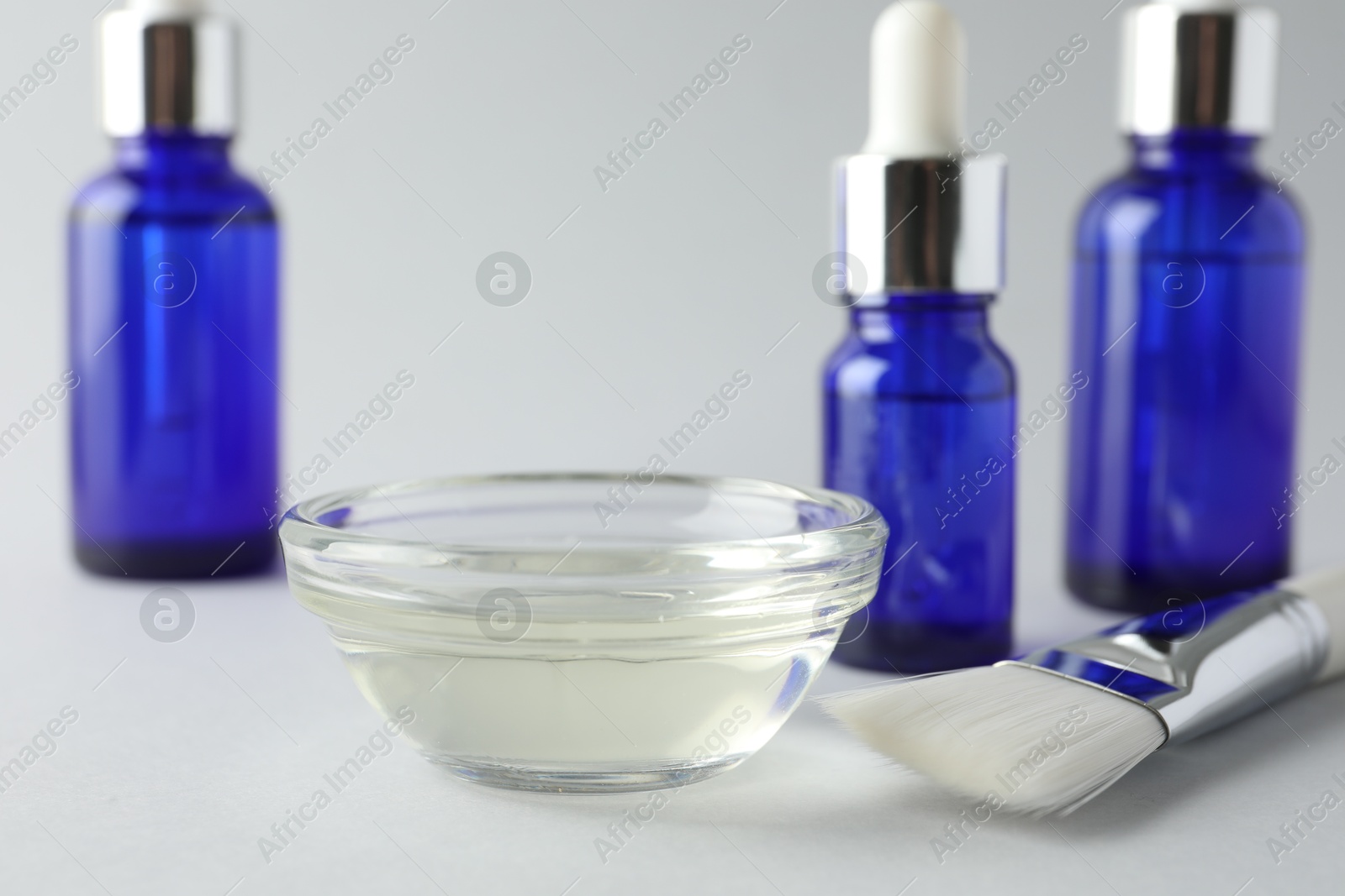 Photo of Chemical peel liquid in bowl, brush and cosmetic bottles on light background, closeup. Peeling procedure