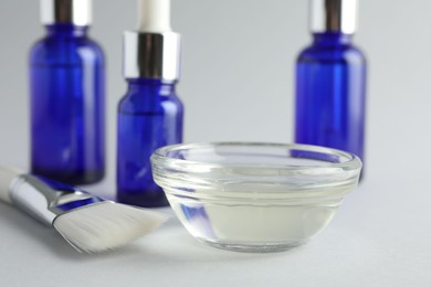 Photo of Chemical peel liquid in bowl, brush and cosmetic bottles on light background, closeup. Peeling procedure