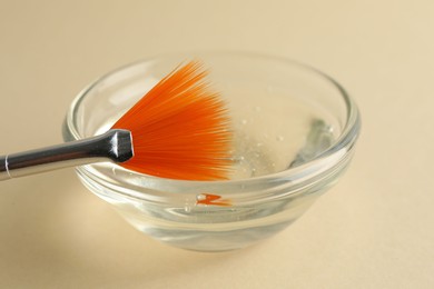 Photo of Chemical peel liquid in bowl and brush on beige background, closeup. Peeling procedure