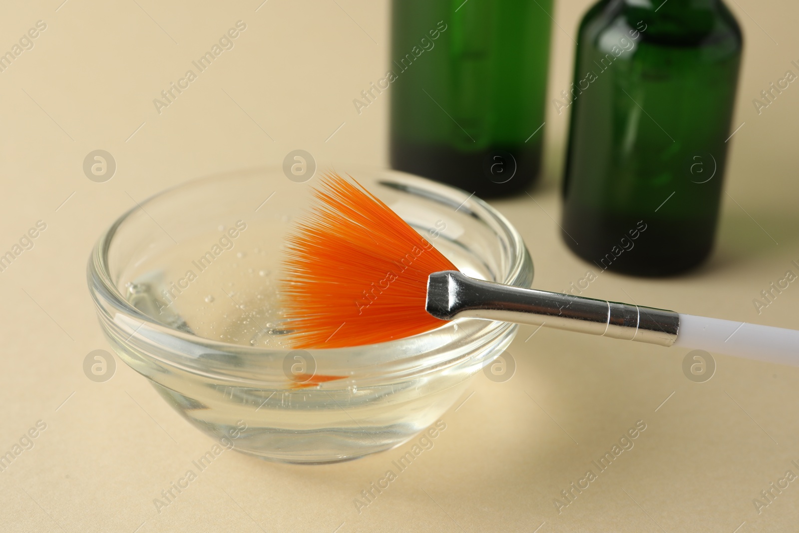 Photo of Chemical peel liquid in bowl, brush and cosmetic bottles on beige background, closeup. Peeling procedure