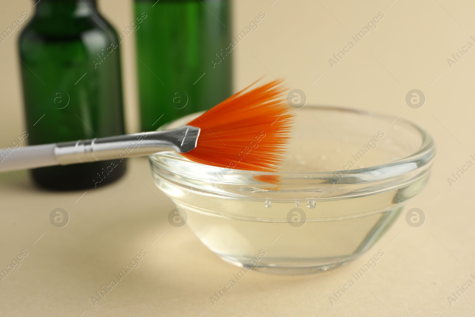 Photo of Chemical peel liquid in bowl, brush and cosmetic bottles on beige background, closeup. Peeling procedure